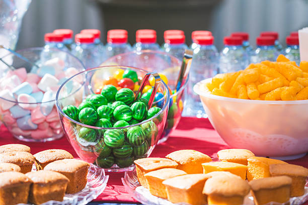 A colorful outdoor party table featuring bite-size cakes, cream cookies, candy, marshmallows, and puffed corn, arranged to create a festive and inviting dessert and snack display in natural daylight.