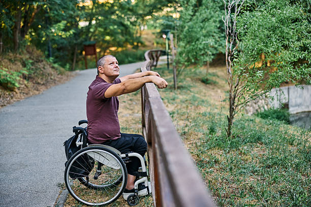 A man in a wheelchair peacefully enjoys the view of nature by a wooden fence.