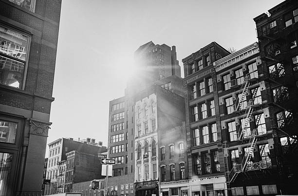 Black-and-white image of a Manhattan street lined with tall brick buildings, sun flare, and a fire escape. Evokes urban rhythm, timeless downtown charm, and cinematic city life.