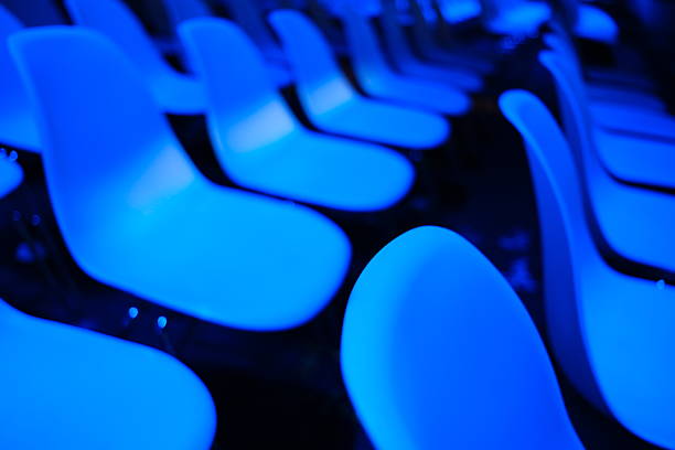 Artistic abstract shot of empty seats in a dark concert hall or conference room. The image features a shallow depth of field with a strong bokeh effect, focusing on the curved shapes of the chairs under vibrant blue stage lighting. Concept of silence, absence, or anticipation