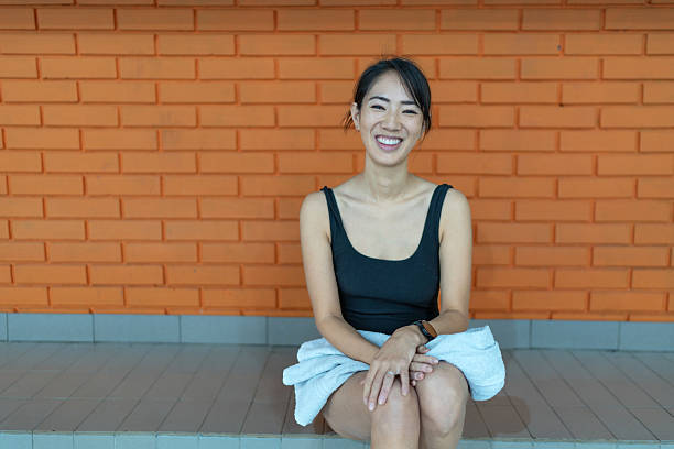 Young asian woman in a black swimsuit smiles confidently at the camera while sitting against an orange brick wall with a white towel on her lap, relaxed and radiant after a workout