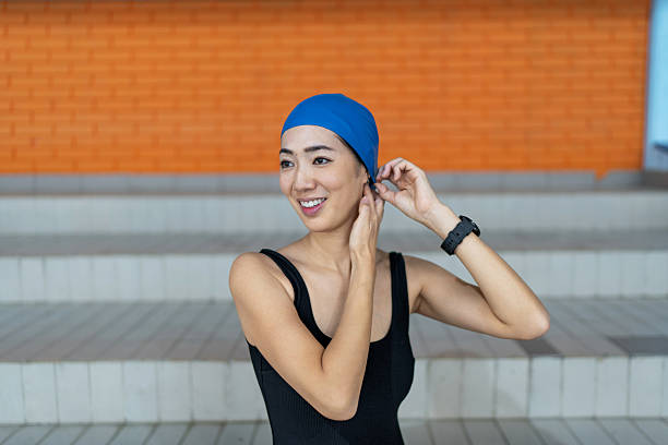 Asian woman wearing a blue swim cap and black swimsuit, preparing for her swimming exercise in a public swimming pool, smiling with happiness and focus on her training