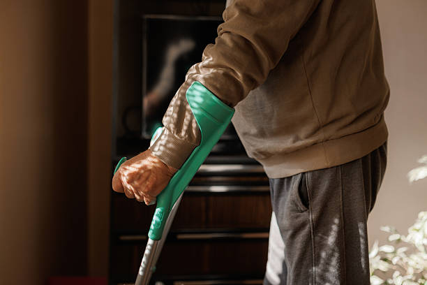 Close-up of an older individual's hand holding a green forearm crutch, with dappled light creating patterns on their skin and clothing