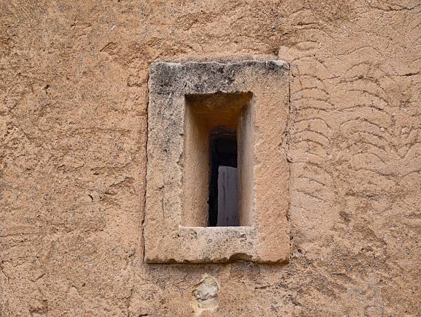 Narrow loophole cutting through plaster wall, slender vertical opening casts deep shadow across rough stucco,
