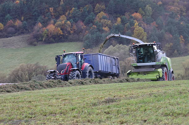 Agricultural machinery harvesting silage on a rural hillside in autumn, with a combine harvester loading chopped grass into a tractor trailer against colorful forested slopes.