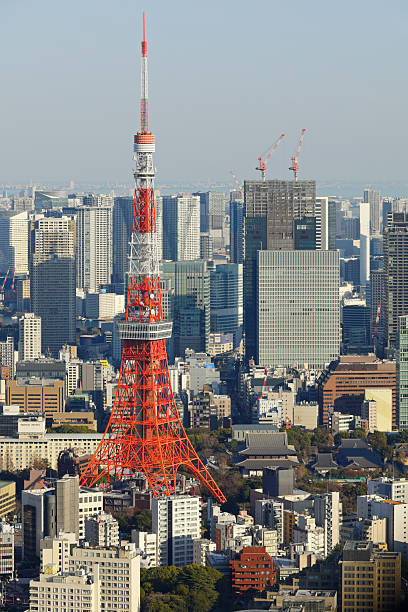 High angle view of the Tokyo skyline in Japan, showing a dense urban area. Tokyo Tower stands prominently at the center of the cityscape, surrounded by dense clusters of modern skyscrapers under a clear daytime sky.