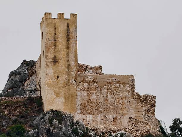 Ancient tower keep rises from rocky base, weathered stone blocks and battlement lines revealing medieval