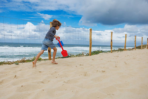 A child runs excitedly on the sandy beach, view from behind holding a red shovel while enjoying a sunny day by the ocean with waves crashing in the background and a wooden fence nearby
