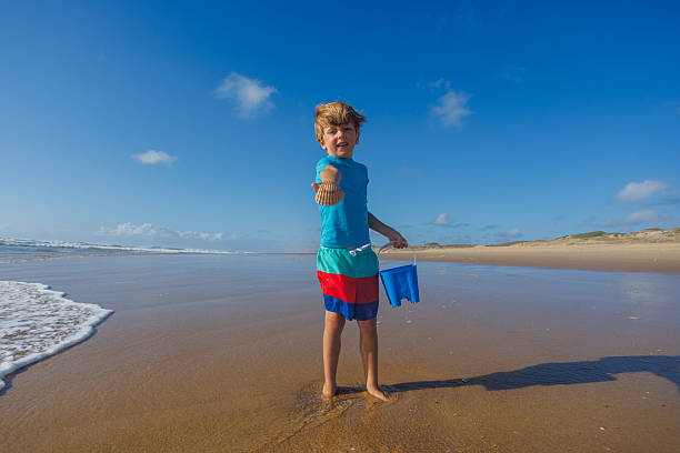 A young boy in swimwear have fun vacations stands on a sandy beach holding a bucket and displaying a seashell, enjoying a sunny day by the ocean with gentle waves