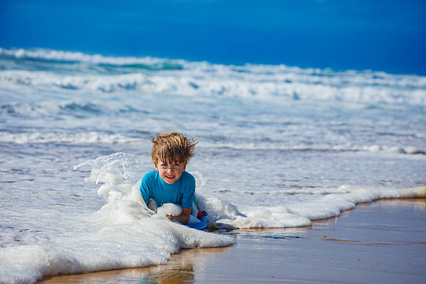 A young boy is joyfully playing in the ocean waves while sitting on the sandy beach, surrounded by foamy water and a bright blue sky in the background