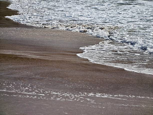dark wet sand with foamy surf, high contrast edge of water and textured froth evoking dramatic coastal mood