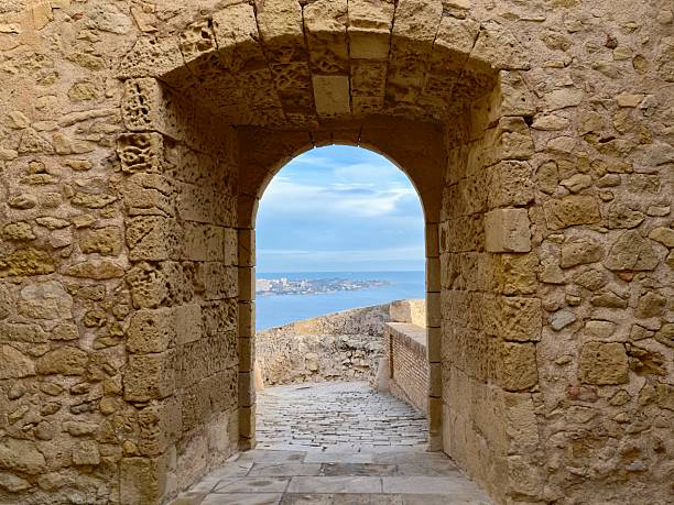 arched stone doorway framing blue sea horizon, coastal walkway leading toward bay, sunlit sky, distant boats,