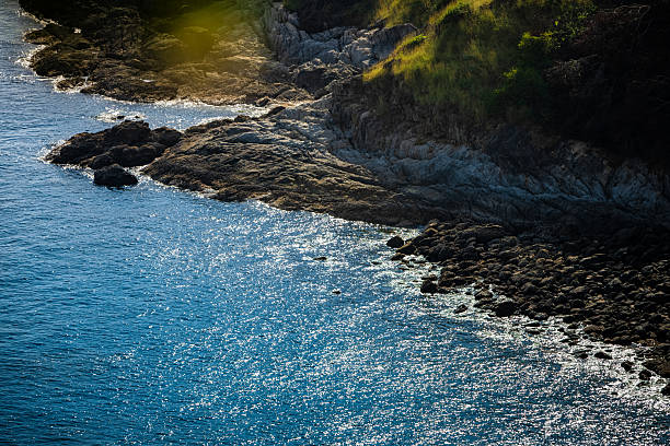 Sunlight reflects on deep blue ocean water along a rugged rocky coastline with natural cliffs and coastal vegetation. Scenic seaside landscape captured in warm natural light without people.