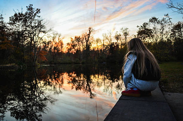Alone girl squatting by water, observing the sunset reflected on the calm pond surface