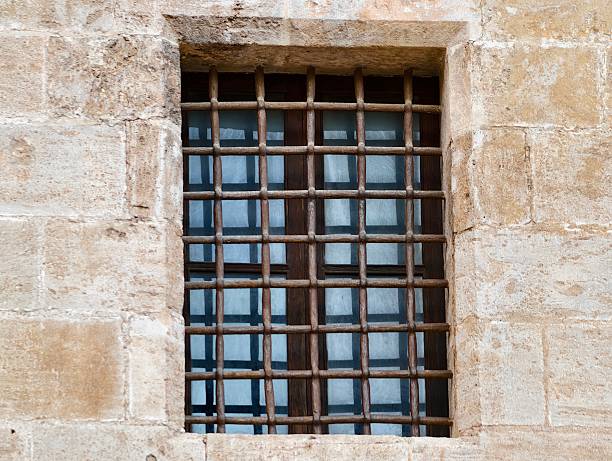 gridded window with reflective glass panes and heavy iron bars set in stone surround, subtle sky reflections,