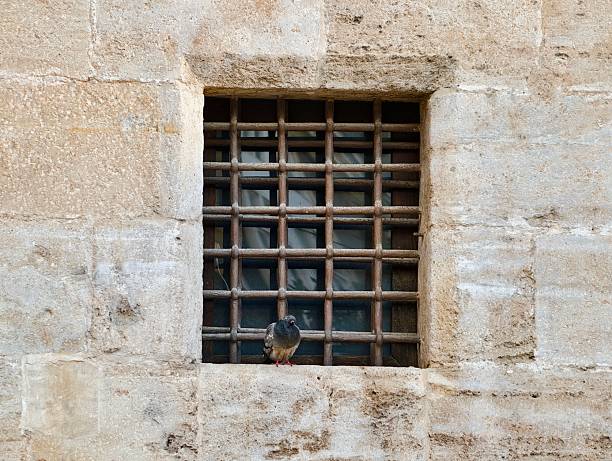 Locked barred window set in stone, visible padlock and chain with heavy shadow and contrast, weathered metal
