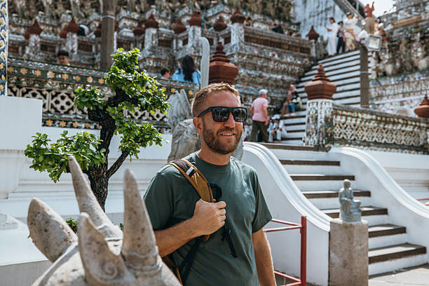Smiling adult man wearing sunglasses and backpack at a colorful historic temple with intricate designs. Male traveler enjoying cultural sightseeing and heritage tourism in Asia. Tourist visiting famous landmark.