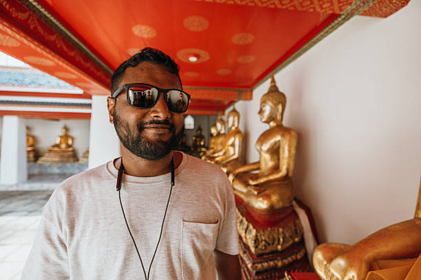 Adult man wearing sunglasses and earphones walking past golden Buddha statues in a temple corridor. Tourist exploring religious site with traditional architecture and cultural artifacts. Spiritual travel and heritage.