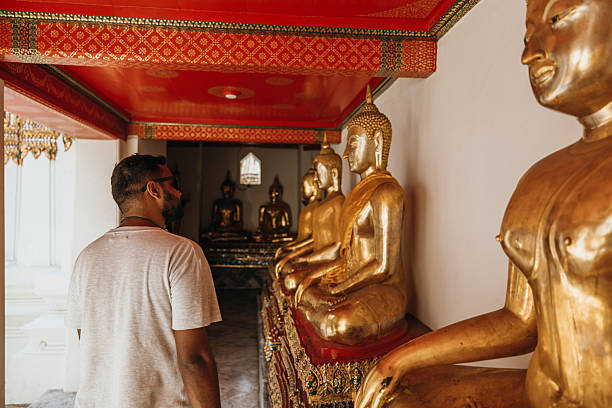 Adult man wearing sunglasses and earphones walking past golden Buddha statues in a temple corridor. Tourist exploring religious site with traditional architecture and cultural artifacts. Spiritual travel and heritage.