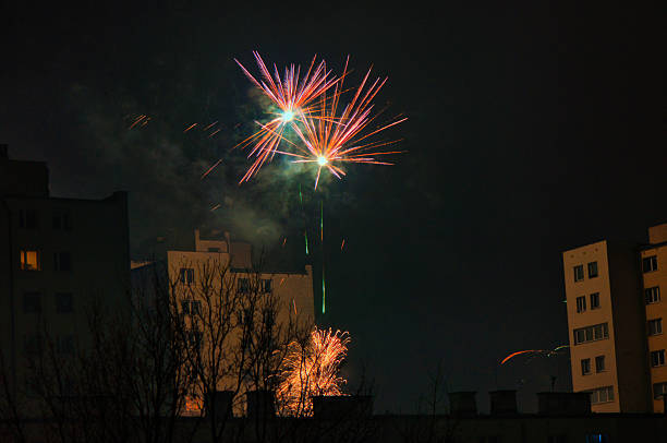 Colorful fireworks explode above a dark skyline, reflecting on windows and silhouettes of apartment blocks. The festive scene conveys excitement, celebration, and shared moments of wonder in an urban night setting.