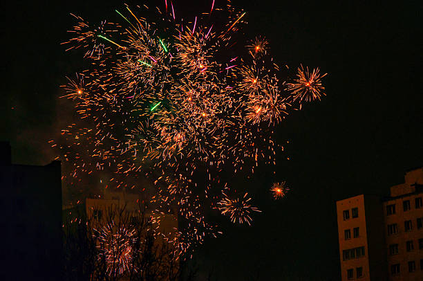 Colorful fireworks explode above a dark skyline, reflecting on windows and silhouettes of apartment blocks. The festive scene conveys excitement, celebration, and shared moments of wonder in an urban night setting.