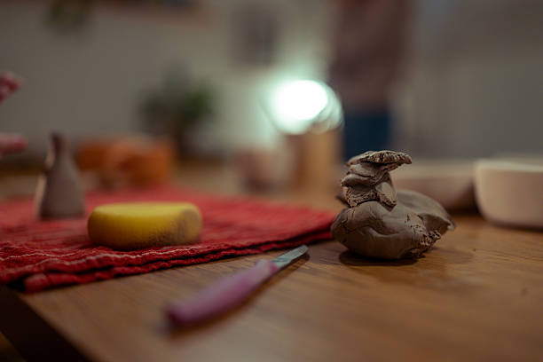 A detailed shot of moist gray clay pieces stacked on a wooden work surface in a pottery studio. In the blurred background, a yellow cleaning sponge rests on a red cloth next to a small sculpting knife with a purple handle
