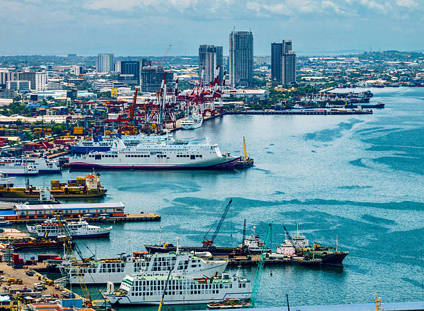 A busy Cebu City port scene with cargo ships, ferries, cranes, and a distant urban skyline. Clear blue water and maritime activity reflect a vibrant Philippine harbor.