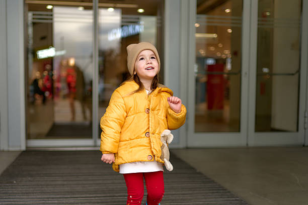 Little girl in yellow jacket and beanie entering a modern shopping mall, looking up with curiosity.