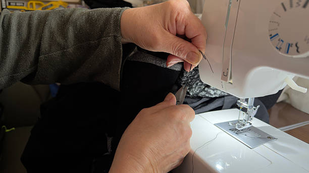 Female shoe factory worker using industrial sewing machine to make detail for new modern black sneakers, needle and hands holding shoe detail in closeup. Footwear manufacturing industry concept