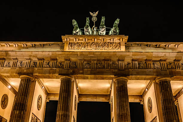 Warm spotlights illuminating the golden stonework of the Brandenburg Gate, iconic symbol of Berlin and Germany reflecting in the cobbles of Pariser Platz under blue dusk skies.