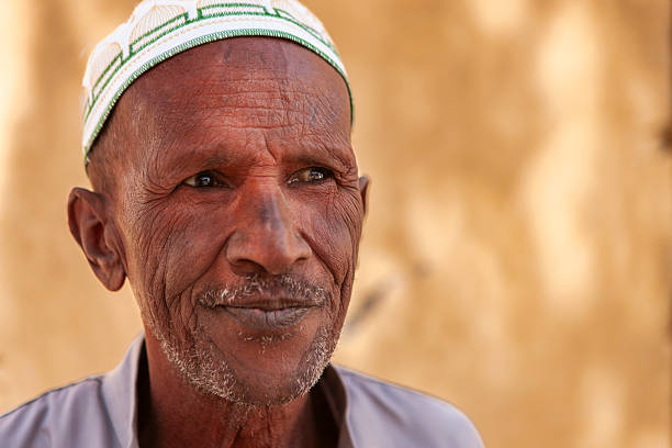 Close-up portrait of an older man with deep facial lines and a gentle, knowing smile. He wears a small white cap with green embroidery and a light-colored shirt; short grey stubble frames his mouth and chin. Warm, soft light and a blurred golden background emphasize the texture of his skin and the quiet, contemplative mood.
