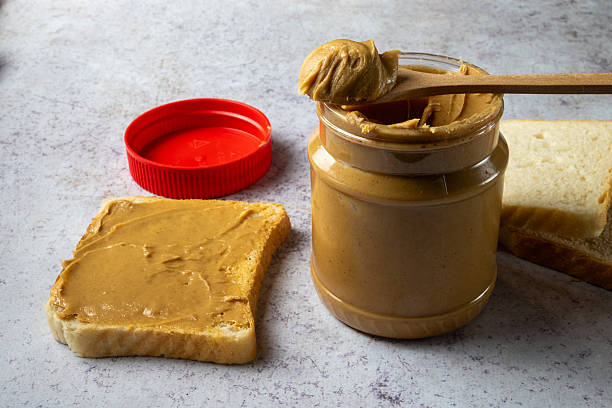 Glass jar of peanut butter with a wooden spoon, sliced white bread, and a slice spread with peanut butter on a neutral kitchen surface.