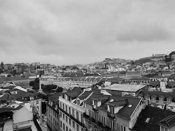 A high-angle, monochrome perspective of the dense rooftops and hilly terrain of Lisbon, Portugal. The image captures the traditional architecture and urban layout of the city under a dramatic overcast sky.