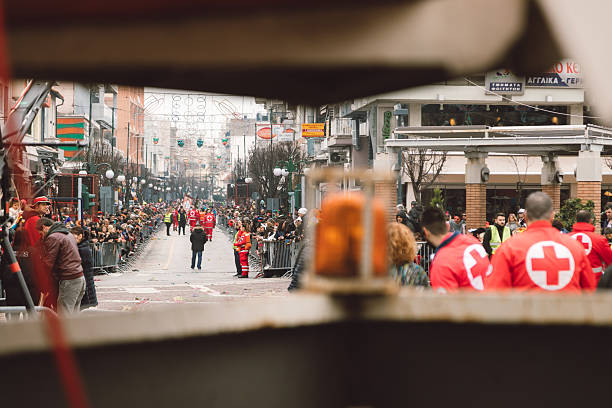 Xanthi, Greece - February 18, 2018: A long perspective view of a crowded city street during the annual Xanthi Carnival is seen through a foreground barrier, with spectators lining both sides of the parade route while Red Cross emergency teams and security personnel stand on duty, emphasizing crowd control, public safety, and the scale of the traditional street celebration in the urban center