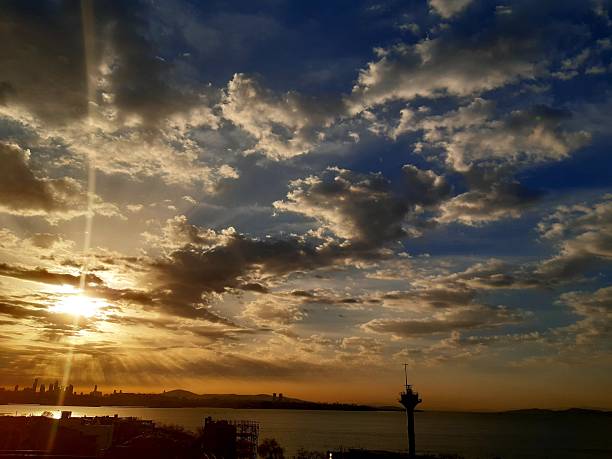 A scenic wide-angle view of a golden sunset over the Bosphorus in Istanbul, featuring dramatic sunbeams through the clouds and the city skyline in silhouette.