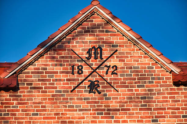Close-up of a red brick facade featuring a black crest-style emblem with crossed lines, a central M/K monogram, and the year 1872, set against a clear blue sky.