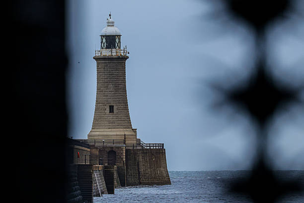 Atmospheric photograph of a historic stone lighthouse standing at the harbour entrance, framed by dark foreground elements and calm sea beyond. This moody coastal scene conveys maritime heritage, navigation, and quiet resilience, making it well suited for travel, editorial, and fine art collections.