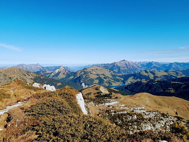 A spectacular panoramic view from a rocky cliff edge in Leysin, showing the distant blue waters of Lac de l'Hongrin nestled between the layers of the Swiss mountain ranges.