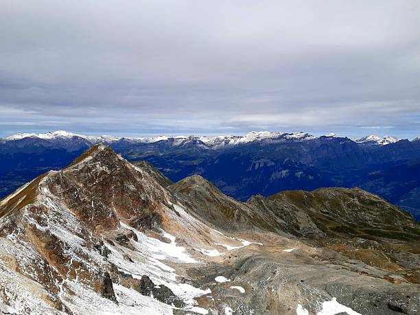 A breathtaking high-altitude vista from the peak of Bella Tola in the Val d'Anniviers, overlooking rugged ridges and distant Alpine ranges under a moody, overcast sky.