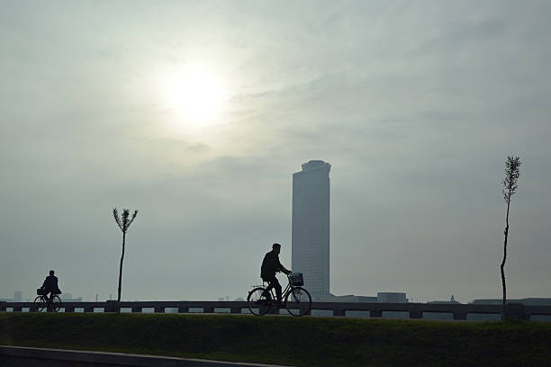 Pyongyang, North Korea - May 1, 2019: local men riding on a bike along Taedong river at sunrise. Modern Yanggakdo Hotel on background