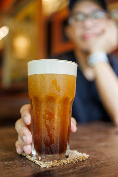 Close-up of a man holding a glass of iced shaken robusta coffee with frothy foam while relaxing at a cozy cafe table. The cold coffee drink is served indoors on a wooden table with natural light, capturing coffee culture, refreshment, and a calm urban lifestyle moment.