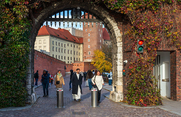Krakow, Poland – October 25, 2025: People walk through a historic arched gate with metal portcullis at the entrance to Wawel Castle. The brick walls and autumn foliage frame daily pedestrian movement in the old city area.