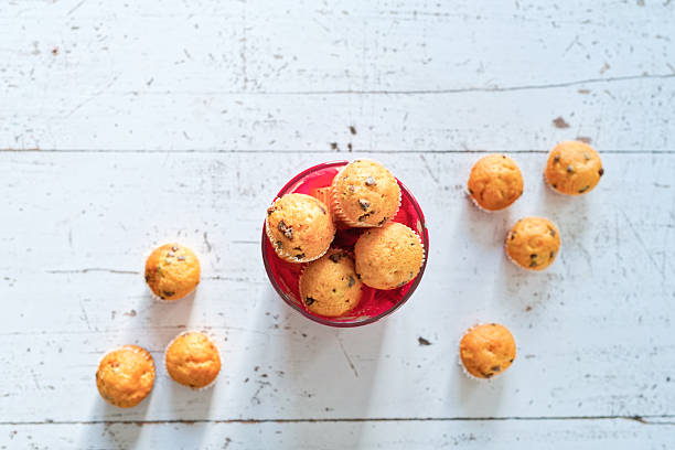 A red cookie jar filled with mini muffins is surrounded by individual muffins scattered across a distressed white wooden table, creating a playful and informal Valentine’s Day snack scene. The bright, airy background provides ample copy space on the left and top for text overlays, banners or social posts, while the golden pastries and long shadows add warmth and a relaxed, homely mood.