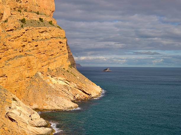 textured sandstone cliff face bathed in warm light overlooking teal ocean, visible rock layers and coastal