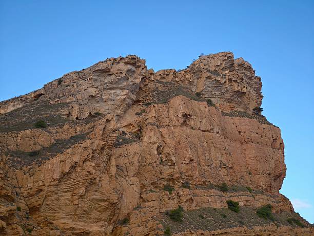 towering rocky cliff against blue sky capturing monumental vertical face, sunlit ledges and sharp shadows