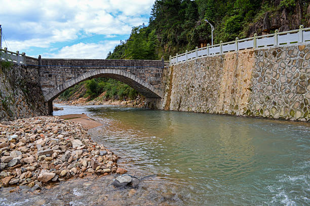 A stone arch bridge spanning a shallow river in a rural mountainous area. The riverbed is filled with rocks and pebbles, and the banks are lined with green trees and a road railing, creating a peaceful countryside scene.