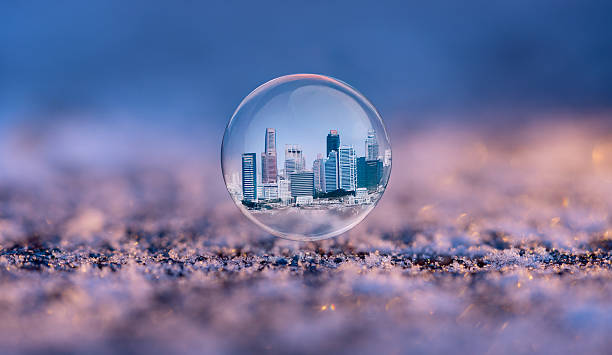 A soap bubble rests on a surface covered with frost. The bubble reflects a city skyline with tall buildings under a clear blue sky. The scene is captured in bright morning light.
