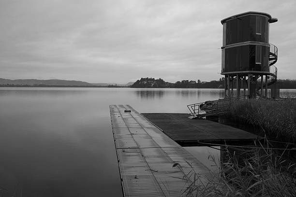 Black and white image of a minimalist lakeside pier leading toward calm water, with a modern structure on the shore. The quiet scene, soft tones, and open space convey stillness, solitude, and balance, creating a strong sense of calm and contemplation.