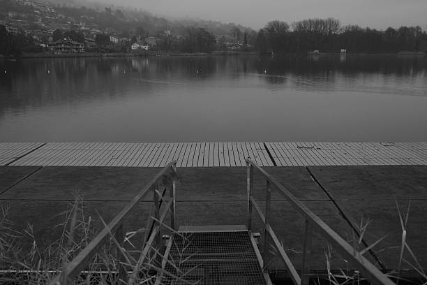 Black and white image of a minimalist lakeside platform leading toward calm water. Strong horizontal lines and still reflections create a quiet, contemplative atmosphere conveying pause, balance, and emotional distance.