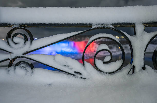 A creative close-up shot of an ornate wrought iron railing heavily covered in fresh white snow. In the soft-focus background, the American Falls at Niagara Falls are illuminated with vibrant blue and red lights, creating a beautiful bokeh effect. This atmospheric night scene captures the quiet, cold beauty of a Canadian winter in Ontario.